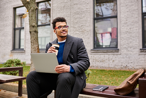 A stylish man with glasses sits on a bench, sipping coffee while typing on his laptop outside.