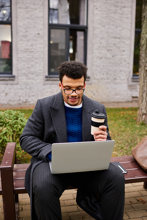 Focused man with glasses types on his laptop, sipping coffee in a tranquil park environment.