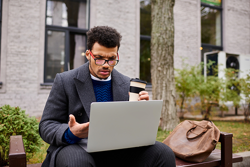 A handsome man enjoys his coffee while working intently on his laptop outdoors.