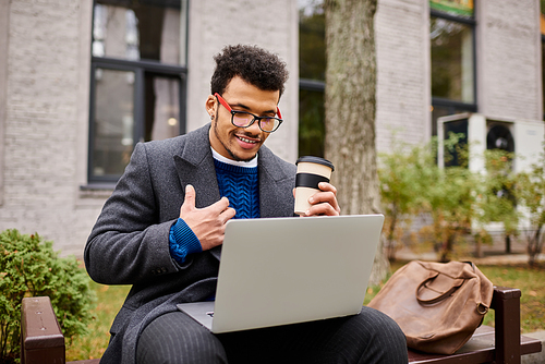 A handsome man casually smiles while sipping coffee and engaging with his laptop outdoors.