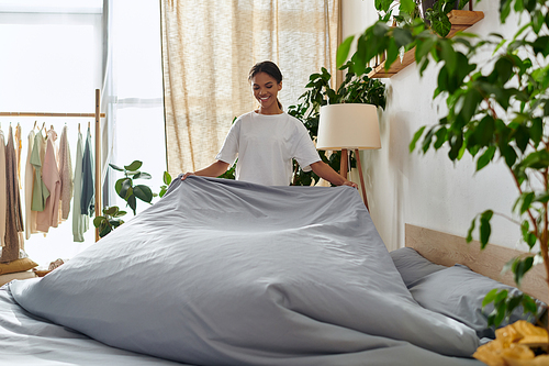 A young African American woman joyfully makes her bed in a stylish and bright apartment.