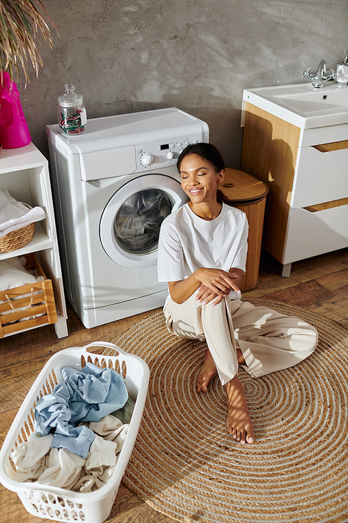 In a modern apartment, a young african american woman relaxes while doing laundry, smiling.