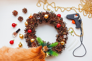 Cute ginger cat near handmade Christmas wreath and things you need to make it - pine cones, glitter, decorative balls and beads, glue gun.