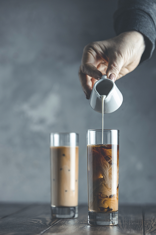 Women hand is pouring homemade sour cream from small jar to glass with brew cold coffee and ice. Cold summer drink on a dark wooden table and gray background with copy space.