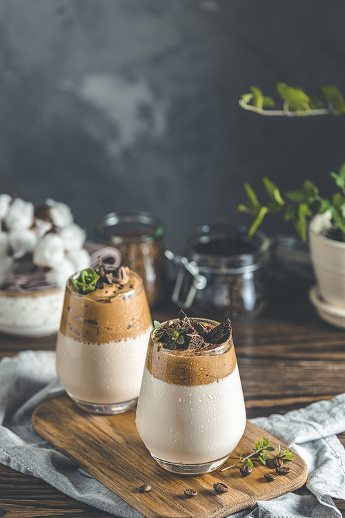 Two glasses of Iced Dalgona Coffee, a trendy fluffy creamy whipped coffee, decorated by thyme and dark chocolate. Dark rustic style.