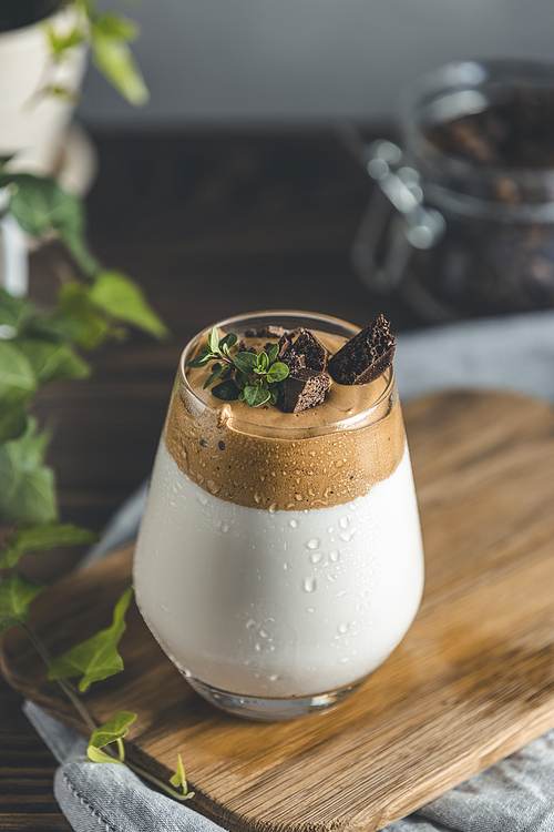 Glass with water drops of Iced Dalgona Coffee, a trendy fluffy creamy whipped coffee, decorated by thyme and dark chocolate. Dark rustic style.