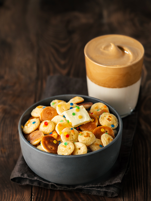 Mini pancakes cereal and dalgona coffee on brown wooden background, copy space. Trendy food and drink - tiny panckakes served sprinkles and whipped instant coffee with milk or dalgona coffee. Vertical
