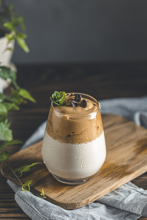 Glass with water drops of Iced Dalgona Coffee, a trendy fluffy creamy whipped coffee, decorated by fresh mint and coffee beans. Dark rustic style.