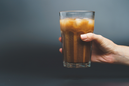 Hand with ice coffee in a tall glass on dark background.