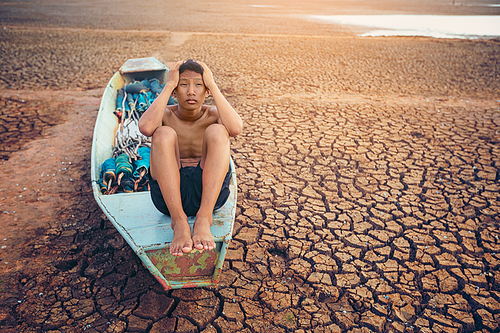 Sad a boy sitting on a boat that was parked in an arid ground for the hope of the sky to rain. Affected of global warming made climate change. Water shortage and drought concept.