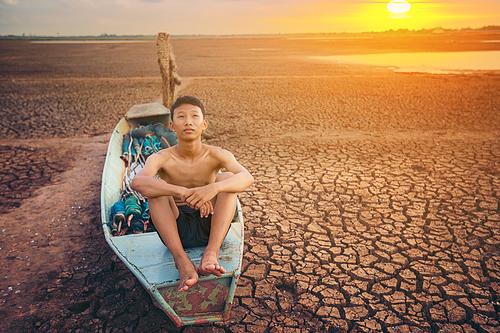 Sad a boy sitting on a boat that was parked in an arid ground for the hope of the sky to rain. Affected of global warming made climate change. Water shortage and drought concept.