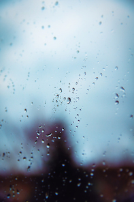 Selective focus of rain drops on a glass window surface with blurry shadows and cloudy background. Dramatic background concept.