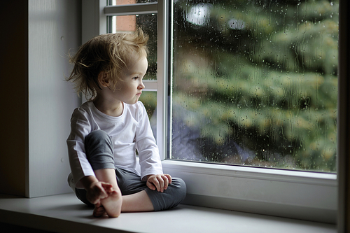 Adorable toddler girl looking at raindrops on the window