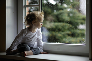 Adorable toddler girl looking at raindrops on the window