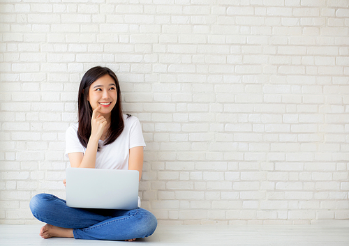 Beautiful of portrait asian young woman working online laptop and thinking sitting on floor brick cement background, freelance girl using notebook computer, business and lifestyle concept.