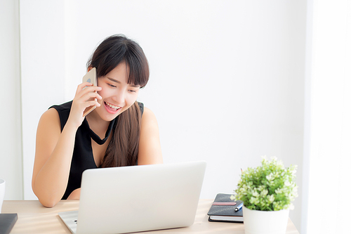 Beautiful young freelance asian woman smiling working on laptop computer and talking mobile smart phone at desk office with professional, girl using notebook, business and lifestyle concept.
