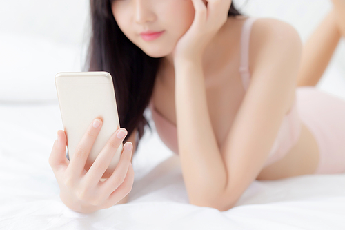Closeup beautiful young asian woman sexy in underwear chatting on mobile phone at bedroom, happy girl in lingerie looking social media on smartphone lying on bed, communication and lifestyle concept.