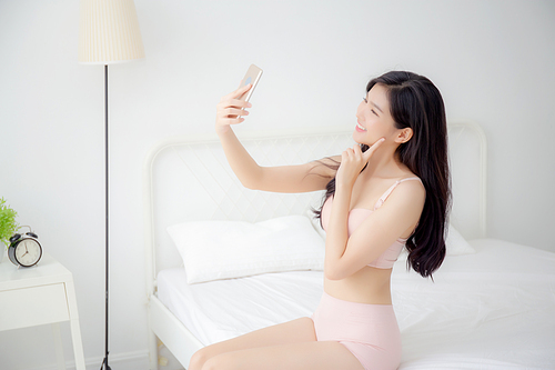 beautiful young asian woman  in underwear talking a selfie on smartphone for social network in the bedroom, girl in lingerie relax with taking a picture on mobile phone on bed in the bedchamber.