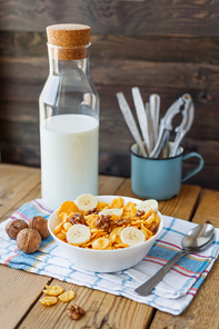 Tasty corn flakes with walnuts and banana in bowl with bottle of milk. Rustic wooden background with plaid napkin. Healthy crispy breakfast snack.