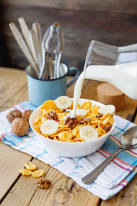 Milk from a bottle flows into the bowl with tasty corn flakes with walnuts and banana. Rustic wooden background with plaid napkin. Healthy crispy breakfast snack.