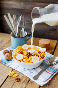 Milk from a bottle flows into the bowl with tasty corn flakes with walnuts and banana. Rustic wooden background with plaid napkin. Healthy crispy breakfast snack.