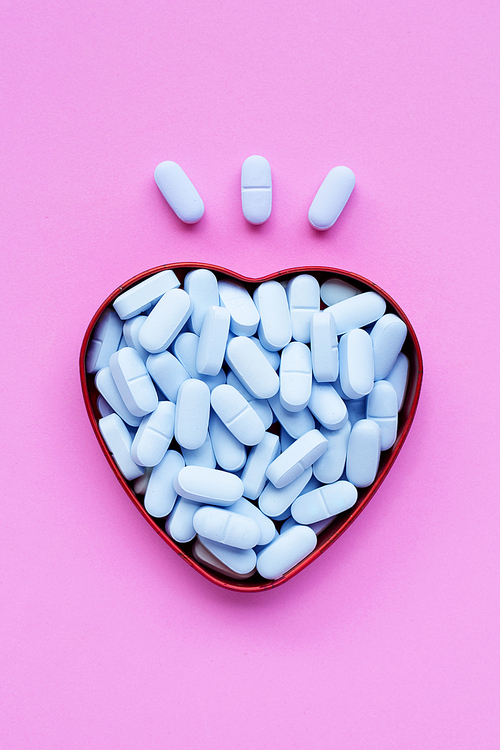 Medical blue pills with heart shaped box on pink background. Top view