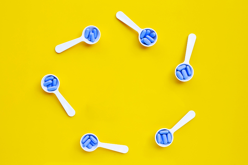 Frame made of medical blue pills with white plastic spoon on yellow background. Top view