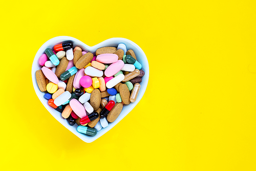 Colorful medicine pills, tablets and capsules in heart shaped bowl on yellow background. Copy space