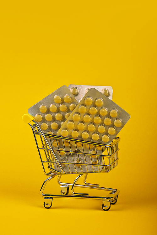 Close up several different blister packs of pills in small shopping cart over yellow background, concept of online medicine order delivery, low angle view