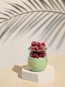 Green matcha overnight oats dressed frozen raspberries. Glass jar with breakfast oats on hexagon pedestal in fashion trendy style with tropical leaf shadow. Modern still life with copy space Vertical