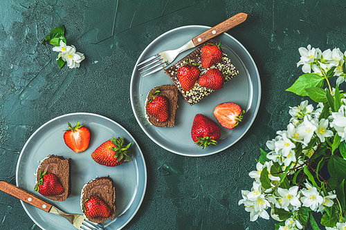 Chocolate rollcake with fresh strawberries in ceramic plate, jasmine and white peonies on dark green concrete surface table. Top view, flat lay, copy space for you text.
