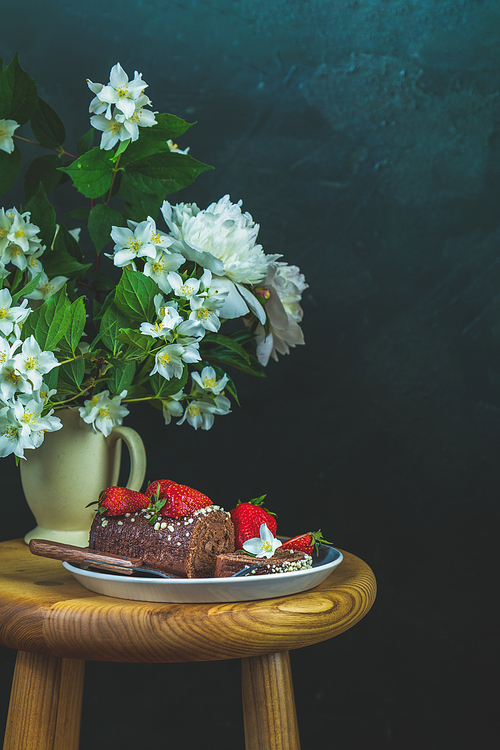 Chocolate roll cake with fresh strawberries, jasmine and white peonies in cup on wooden table. Artistic Still Life in the style of Dutch painting. Copy space for you text..