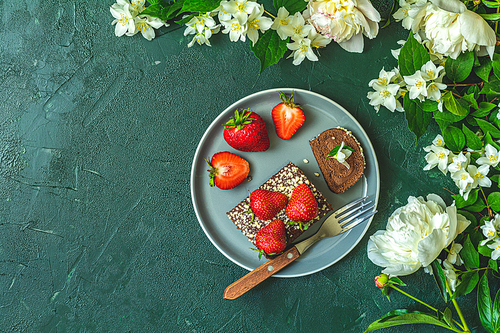 Chocolate rollcake with fresh strawberries in ceramic plate, jasmine and white peonies on dark green concrete surface table. Top view, flat lay, copy space for you text.