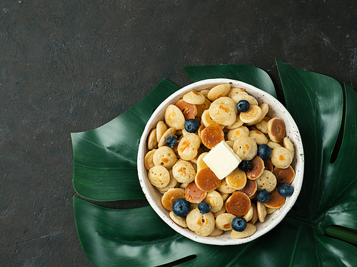 Trendy food - pancakes cereal served butter and blueberries. Heap of mini cereal pancakes in boul on tropical monsterra leaf over dark background. Copy space right for text. Top view or flat lay.