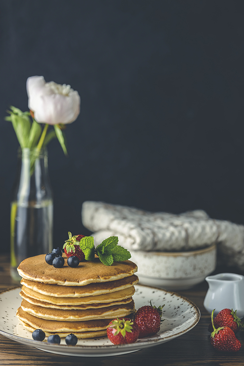 Pancake with strawberry, blueberry and mint in ceramic dish, syrup from small ceramic jar and flowers on a dark wooden table and black background.