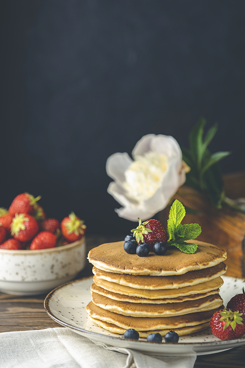 Pancake with srtawberry, blueberry and mint in ceramic dish, syrup from small ceramic jar and flowers on a dark wooden table and black background.