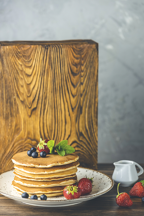 Pancake with srtawberry, blueberry and mint in ceramic dish, syrup from small ceramic jar on a light wooden table and gray background.