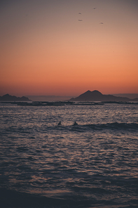 Two surfers in the sea in front of the islands during a sunset