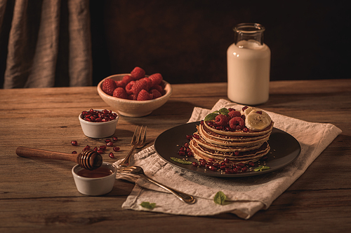 Pancakes with raspberries, banana slices, pomegranate seeds and honey on wooden vintage table.