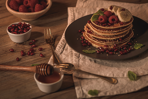 Pancakes with raspberries, banana slices, pomegranate seeds and honey on wooden vintage table.