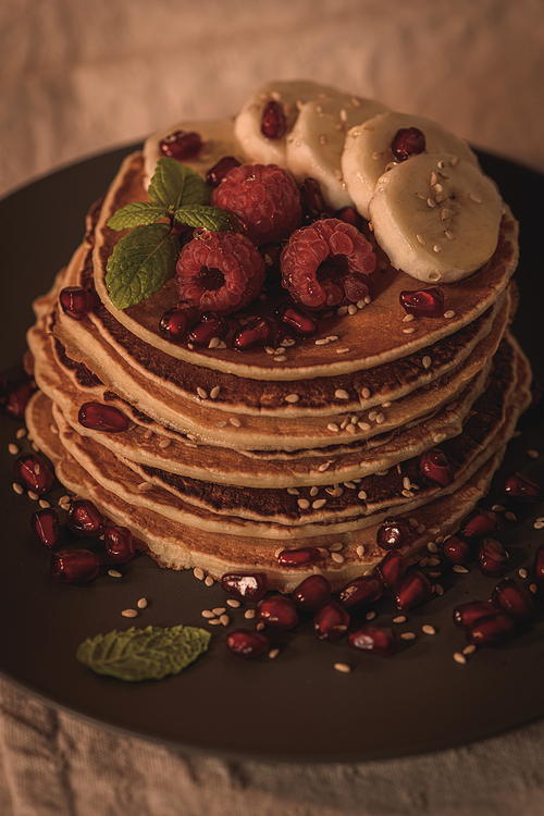 Pancakes with raspberries, banana slices, pomegranate seeds and honey on wooden vintage table.