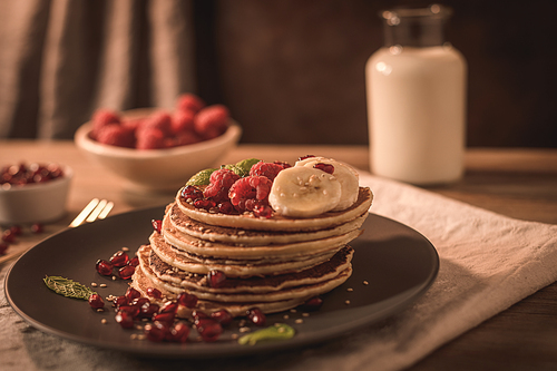 Pancakes with raspberries, banana slices, pomegranate seeds and honey on wooden vintage table.