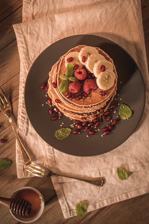 Pancakes with raspberries, banana slices, pomegranate seeds and honey on wooden vintage table.