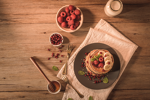 Pancakes with raspberries, banana slices, pomegranate seeds and honey on wooden vintage table.