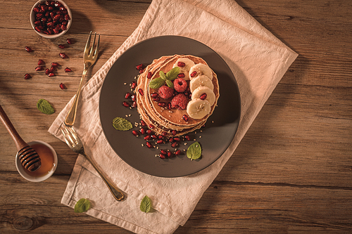 Pancakes with raspberries, banana slices, pomegranate seeds and honey on wooden vintage table.