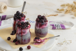 Chia pudding with blackberries, three portions in glass jars on a white table.