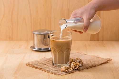 Pouring milk in to glass of coffee on a wooden table.