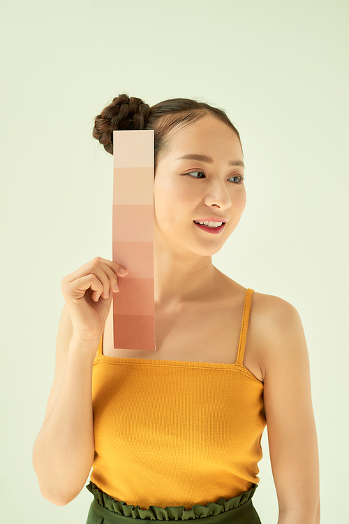 Smiling young woman with skin tone palette or samples while standing over light background