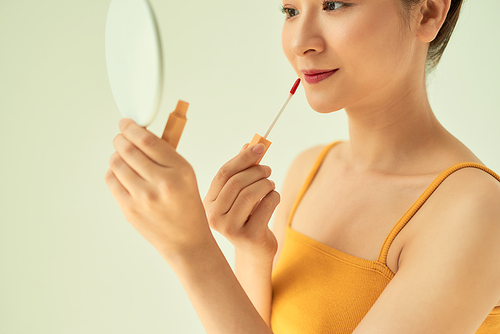 Closeup of Asian young female applying lipstick with mirror against light background.