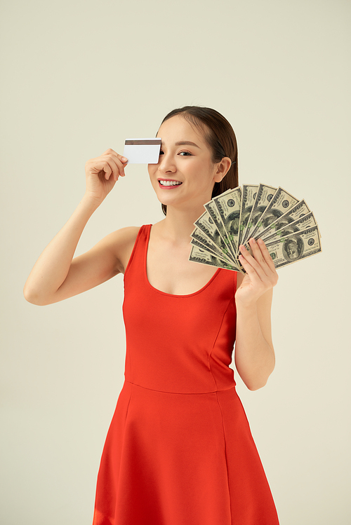 Image of young asian woman holding money banknotes and credit card isolated over gray background